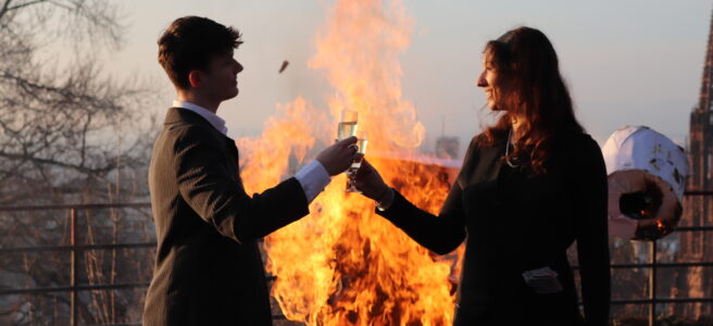 Zwei Menschen in Businesskleidung stoßen mit Sektgläsern an, im Hintergrund brennt eine "1,5 °C" Skulptur